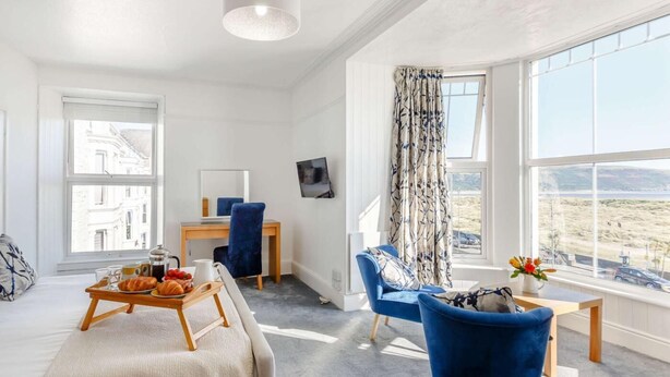 A modern room with a sofa, a dining table with a tray of food, a chair, a small table with flowers, and a large window with a view. - 8 Bed in Barmouth (oc-89784) (Barmouth)