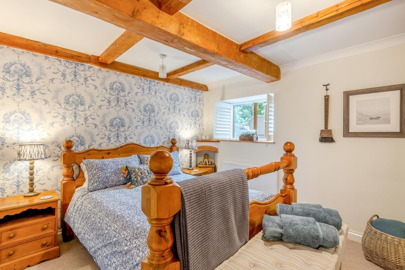 Un dormitorio con una cama de madera, una mesita de noche, una lámpara, una ventana con contraventanas, un marco de cuadro y una escoba.