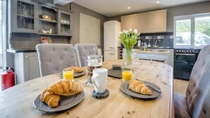 Una tavola imbandita per la colazione con croissant, succo di frutta e una caffettiera, in una cucina moderna con sedie grigie e un vaso di fiori.