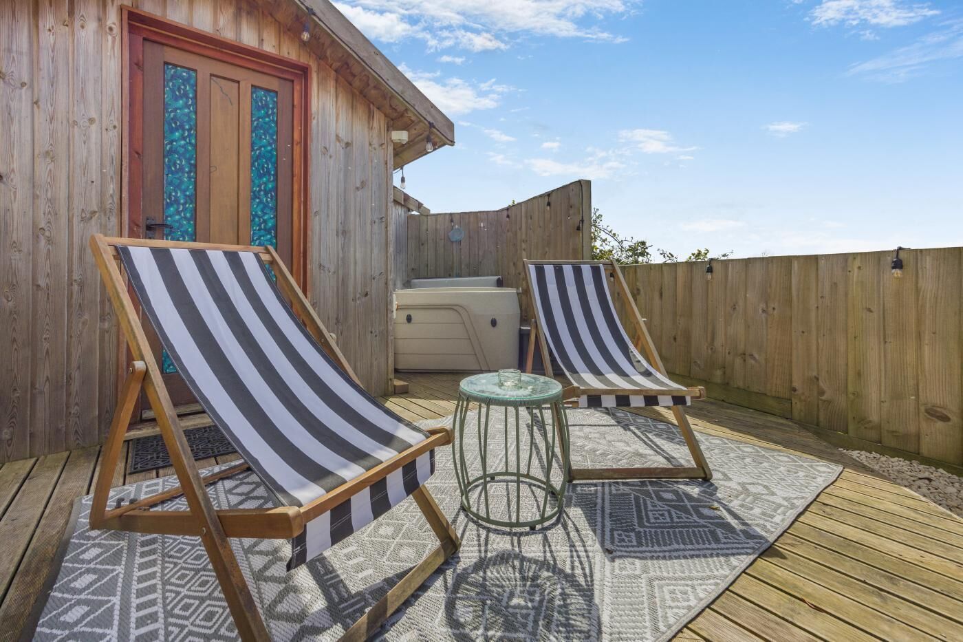 A wooden deck with two striped lounge chairs, a small round table, and a hot tub.