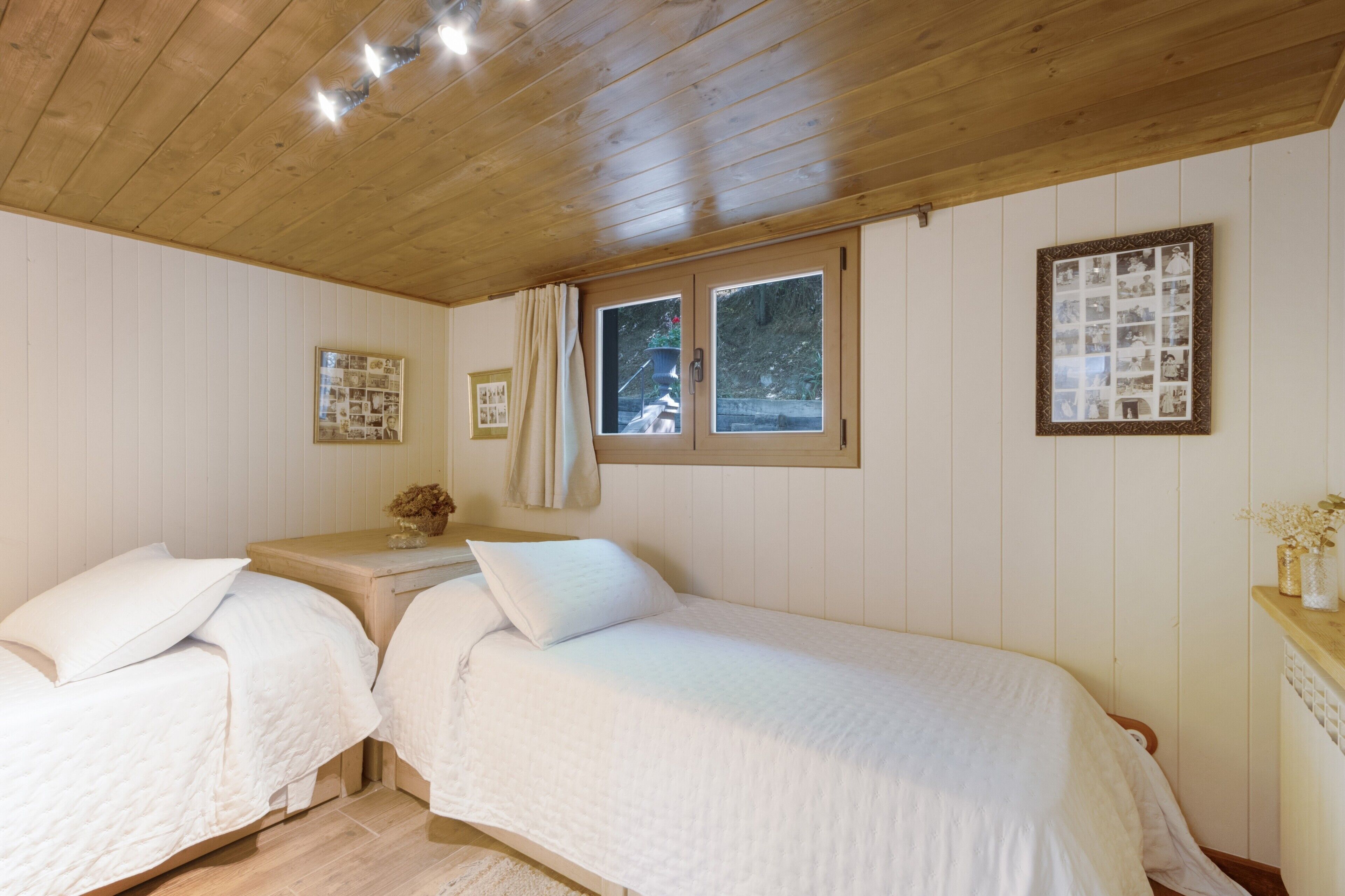 A room with two beds, wooden ceiling, white walls, a window with curtains, and a framed photo on the wall.