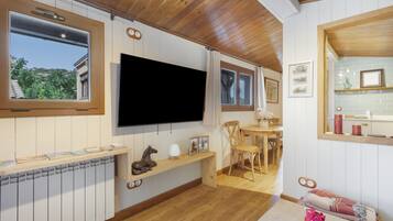 Une pièce dotée d’un plafond en bois, d’un téléviseur à écran plat fixé au mur, d’un banc en bois orné d’une sculpture de cheval, d’un coin repas avec une table et des chaises, et d’une fenêtre donnant sur un paysage verdoyant.