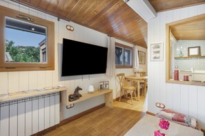 Habitación con techo de madera, televisor de pantalla plana montado, banca de madera con escultura de caballo, zona de comedor con mesa y sillas, y ventana con vista a follaje.