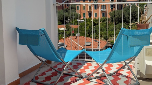 Un balcon avec deux chaises bleues, un tapis à motifs et une vue sur des bâtiments en arrière-plan.