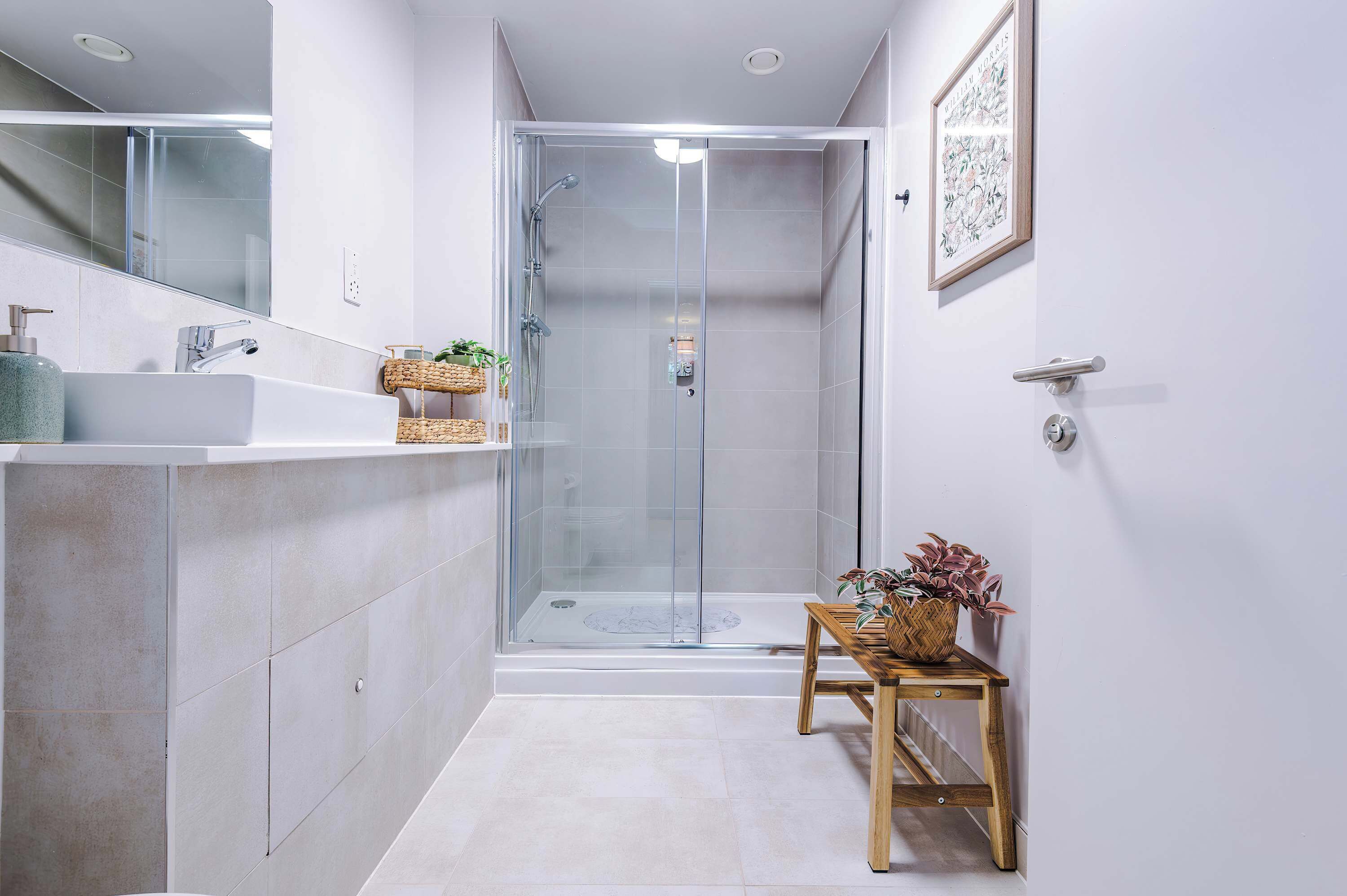 Een moderne badkamer met een douchecabine van glas, een wastafel met spiegel en een klein houten tafeltje met een plant.