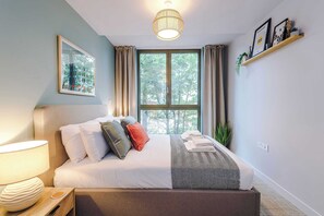 Ein ordentlich bezogenes Bett mit Kissen, eine Nachttischlampe, ein Fenster mit Vorhängen, eine Wandablage mit Dekoration und ein gerahmtes Bild.