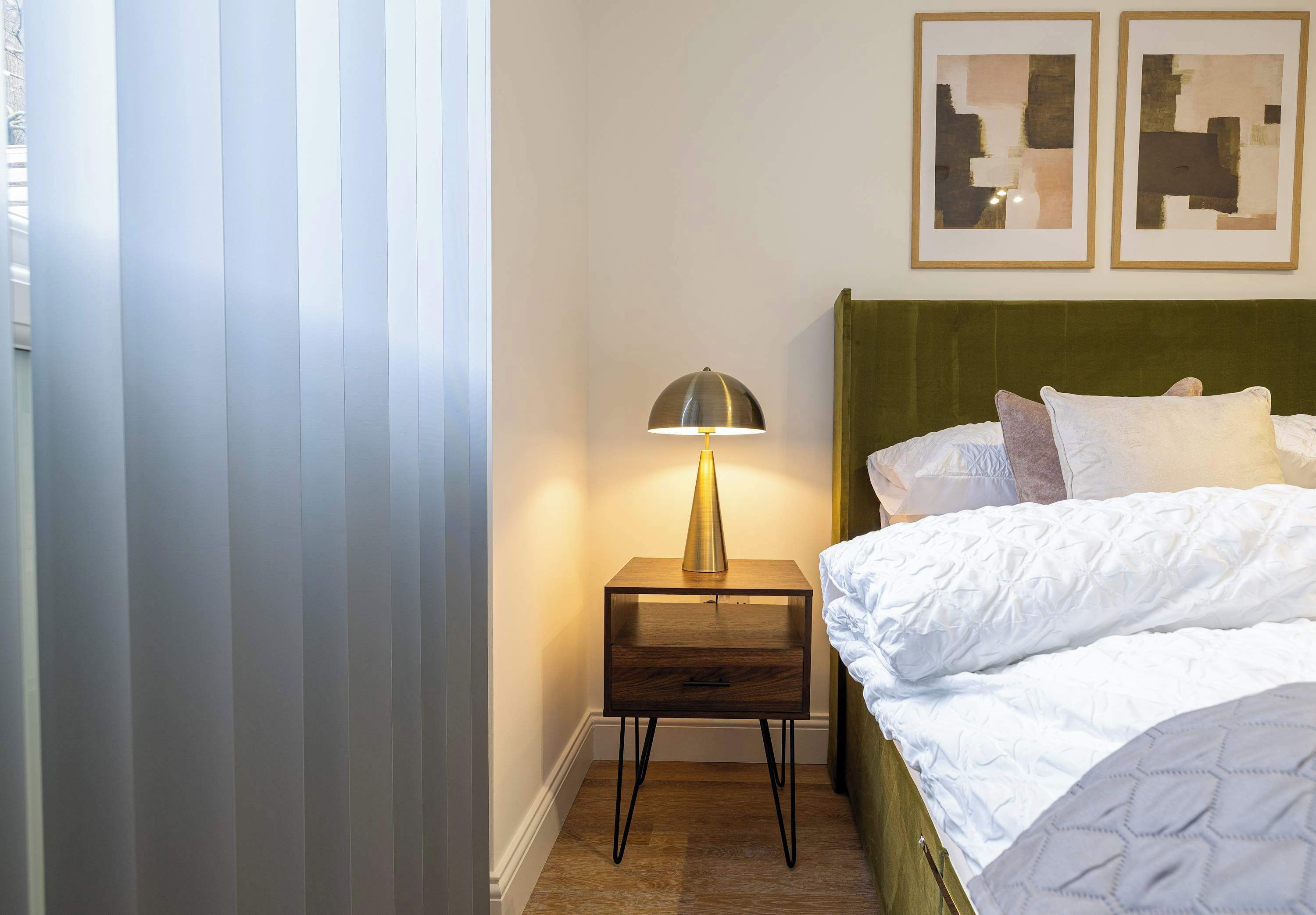Un lit bien fait, avec une tête de lit verte, une lampe de chevet et une œuvre d’art abstraite accrochée au mur.