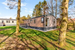 A modern single-story mobile home with a deck and a small porch, surrounded by trees and a grassy yard. - 2 Bed in Pocklington (oc-s39067) (Pocklington)