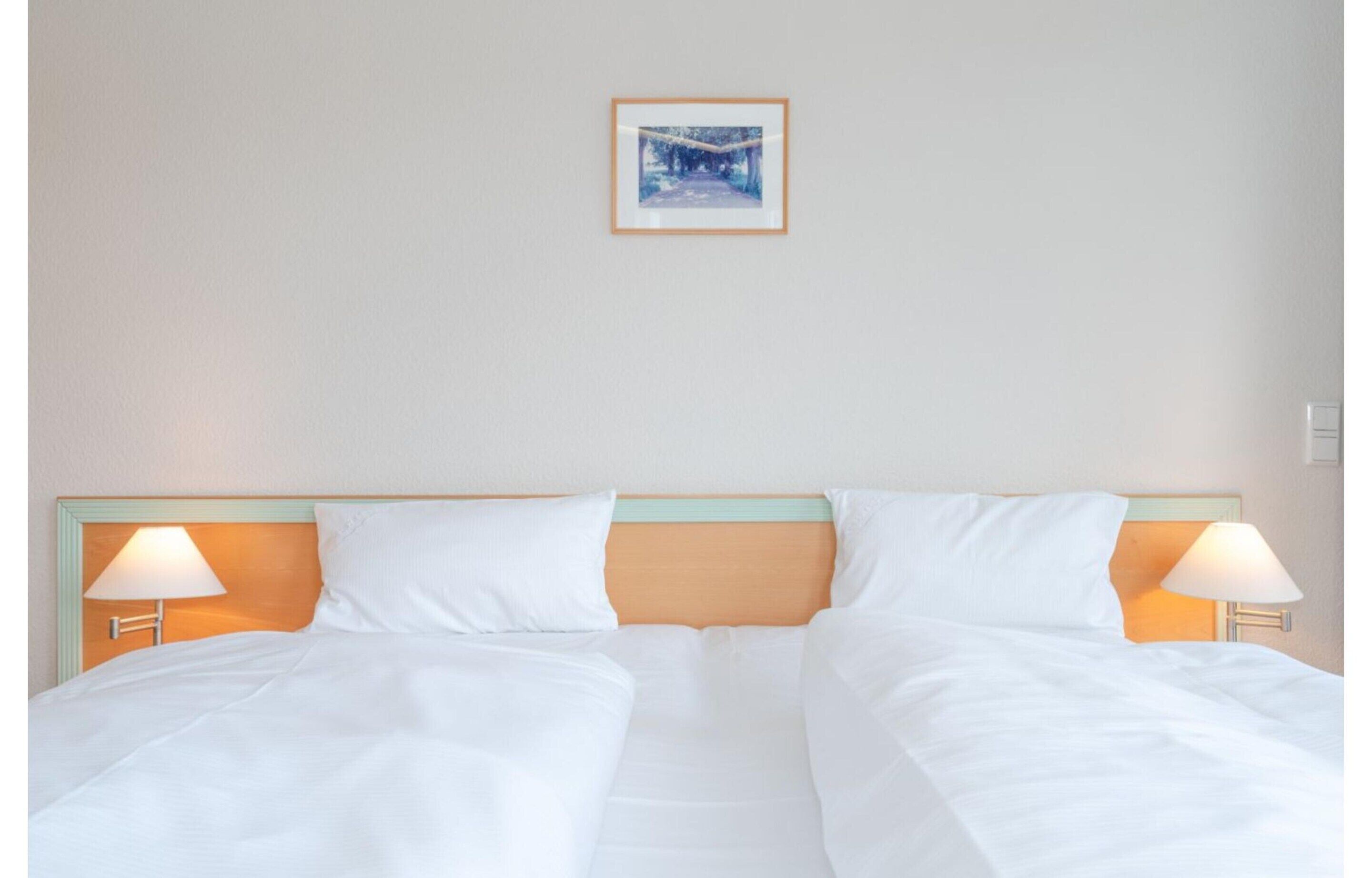 Ein Doppelbett mit weißen Laken, zwei Nachttischlampen und ein gerahmtes Bild an der Wand.