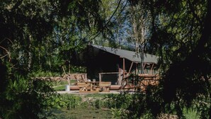 A rustic cabin with a wooden deck, surrounded by dense foliage and a pond. - 3 Bed in Hintlesham (oc-r39060) (Hintlesham)