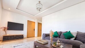 Een moderne woonkamer met een flatscreen televisie, een houten tv-meubel, een grijze bank met groene kussens, een salontafel met decoratieve objecten, en een kroonluchter.