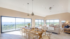 A spacious living area with a dining table, chairs, and a sofa. Large windows offer a view of the landscape. - 5 Bed in Mynytho (oc-t39070) (Mynytho)