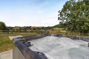 A hot tub with a wooden deck, surrounded by a wooden fence, set against a rural landscape with hills and trees.