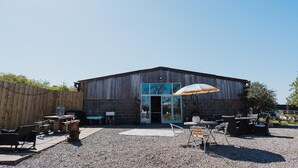 Un bâtiment à l’extérieur en bois, avec une porte d’entrée en verre et un espace extérieur aménagé avec des tables et des chaises.