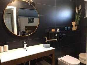 Ein modernes Badezimmer mit rundem Spiegel, weißem Waschbecken, Toilette und dunkel gefliesten Wänden.