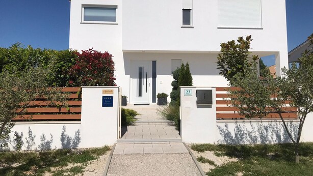 A modern two-story building with a white facade, a central entrance, and a small garden area with shrubs and a bench. - Comfortable Family Retreat With Pool (Vodnjan)