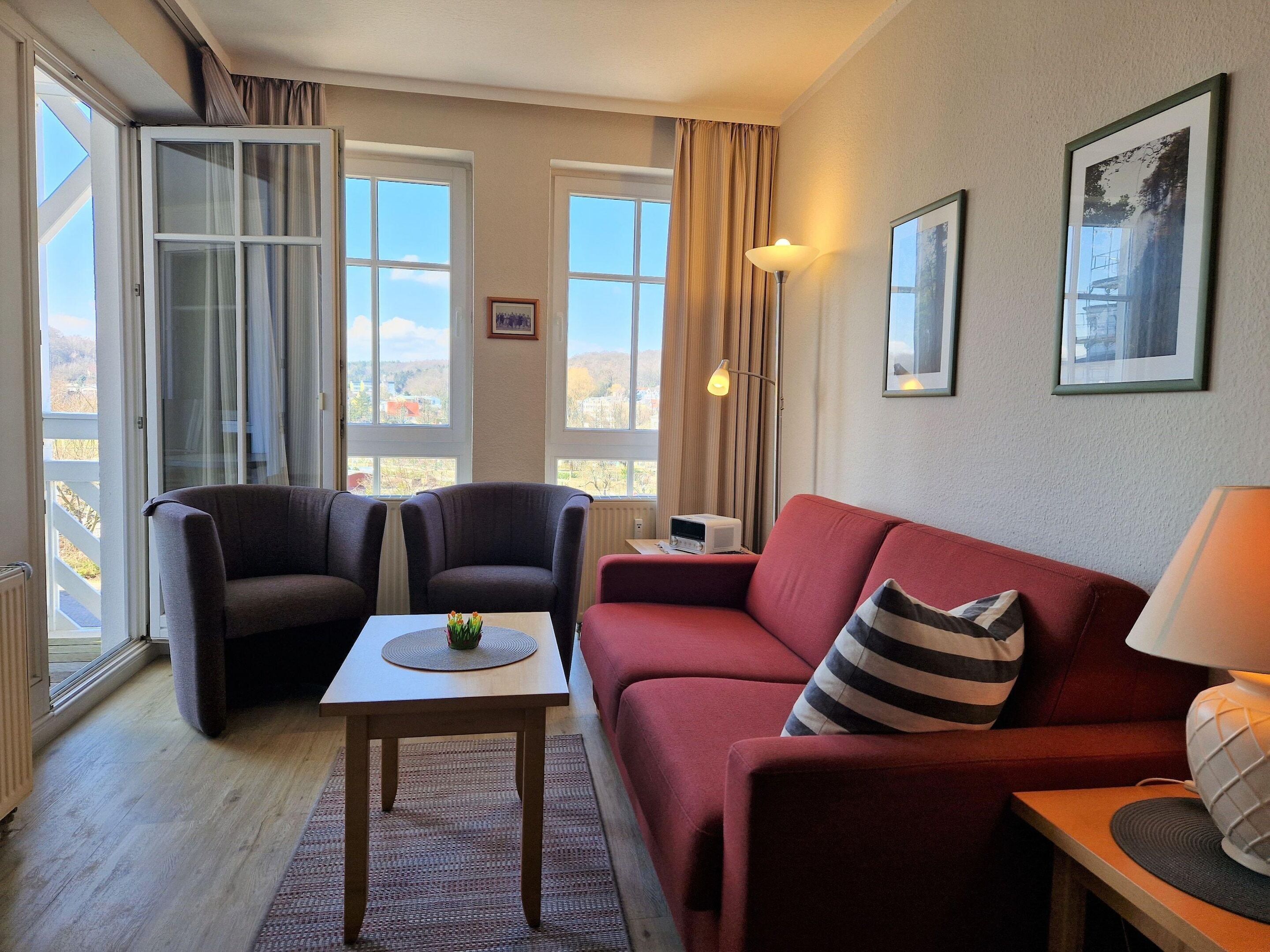 Un salon avec un canapé rouge, deux fauteuils, une petite table sur laquelle se trouve un vase, et une lampe. Il y a deux fenêtres avec des rideaux, un tableau encadré au mur et un radiateur.