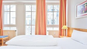 A hotel room with a large bed, a chair, a nightstand with a lamp, and a view of buildings through the windows. - Komfortable Ferienwohnung fur bis zu 4 Personen (Sellin)