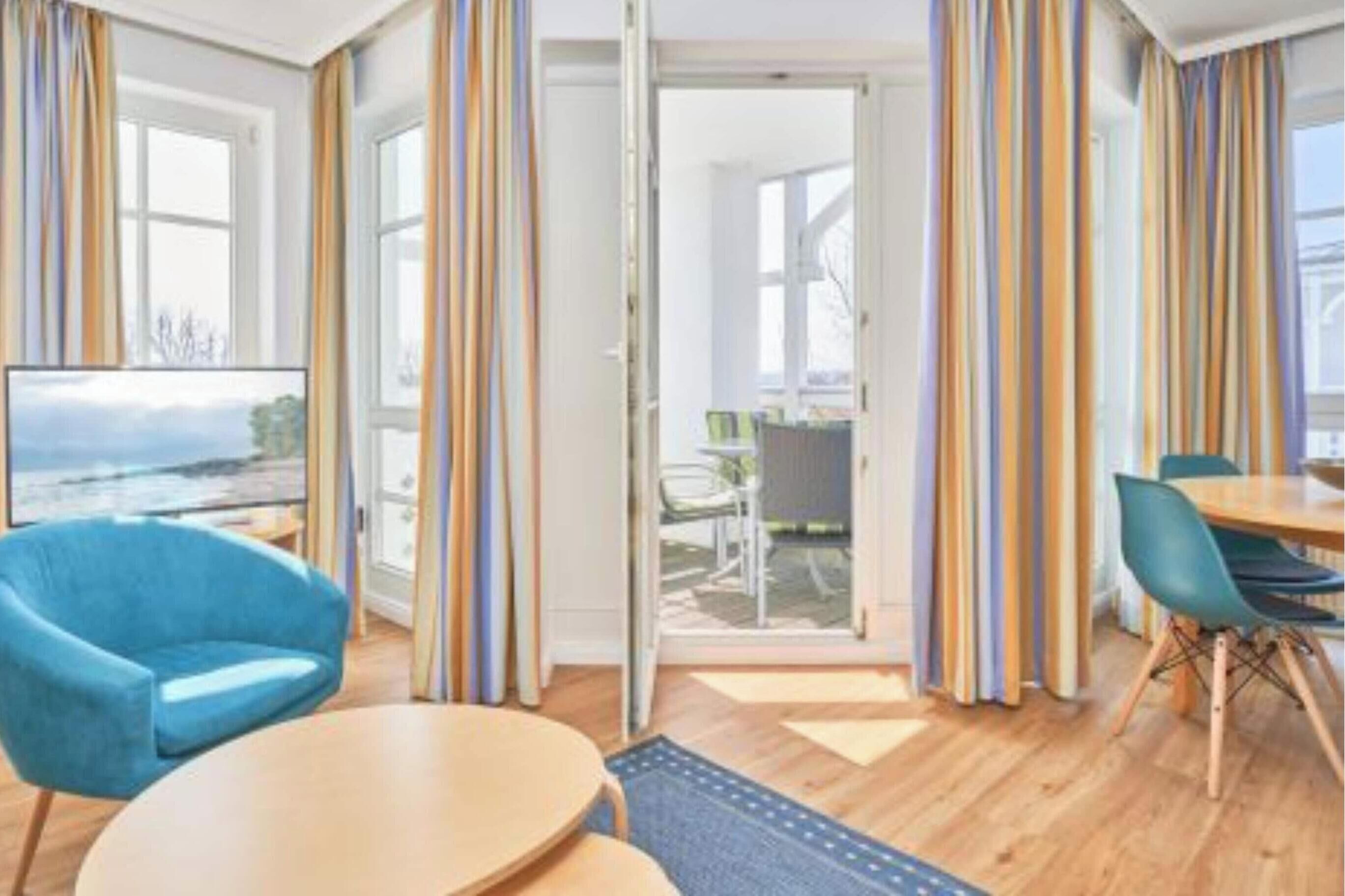 Quarto de hotel moderno com uma grande janela, uma poltrona azul, uma mesa redonda e uma área de refeições com cadeiras.