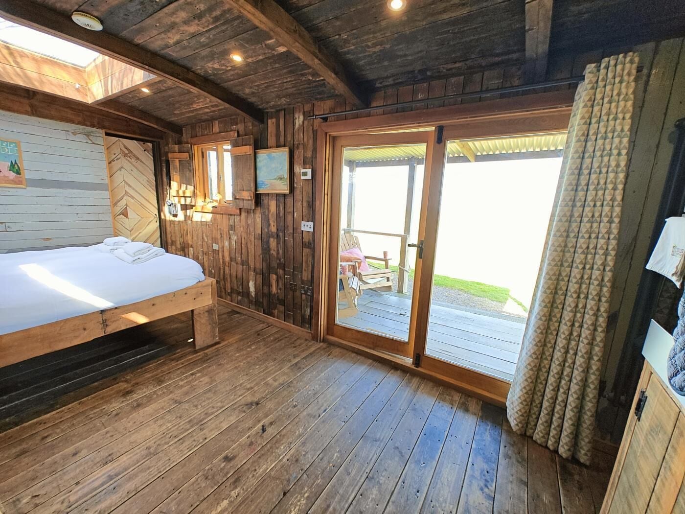 A wooden cabin room with a bed, a chair, and a sliding glass door leading to an outdoor area.