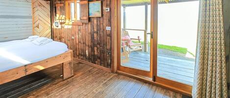 Habitación tipo cabaña de madera, con cama, techo de madera, puerta que da a una terraza exterior y una pequeña mesa con una silla.