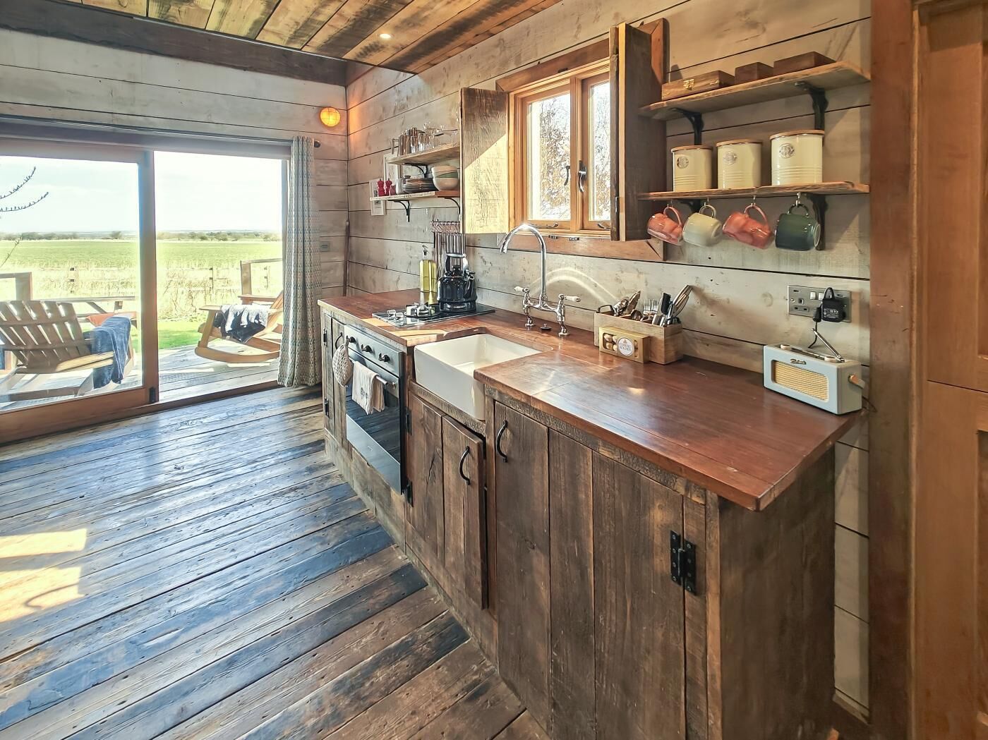 A rustic kitchen with wooden cabinets, a built-in oven, a sink, and a window overlooking a deck.
