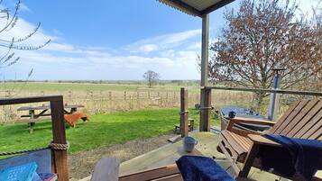 A covered outdoor seating area with wooden benches and a picnic table, overlooking a grassy field and a tree.