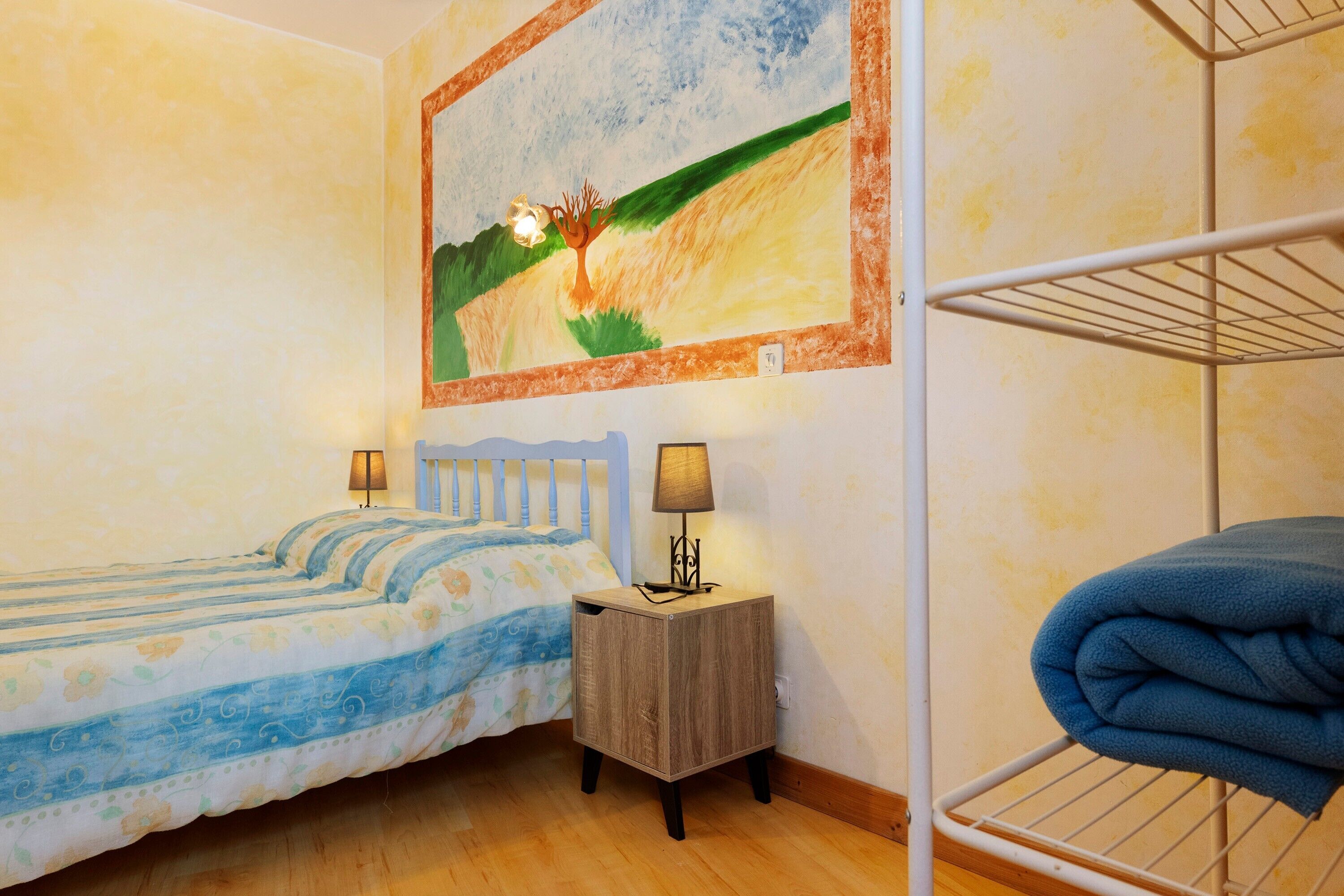 Une chambre avec deux lits, une petite table de chevet avec des lampes, une étagère fixée au mur et un tableau représentant un paysage.