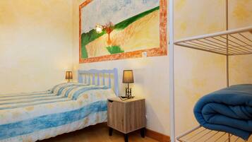 Une chambre avec deux lits, une petite table de chevet avec des lampes, une étagère fixée au mur et un tableau représentant un paysage.