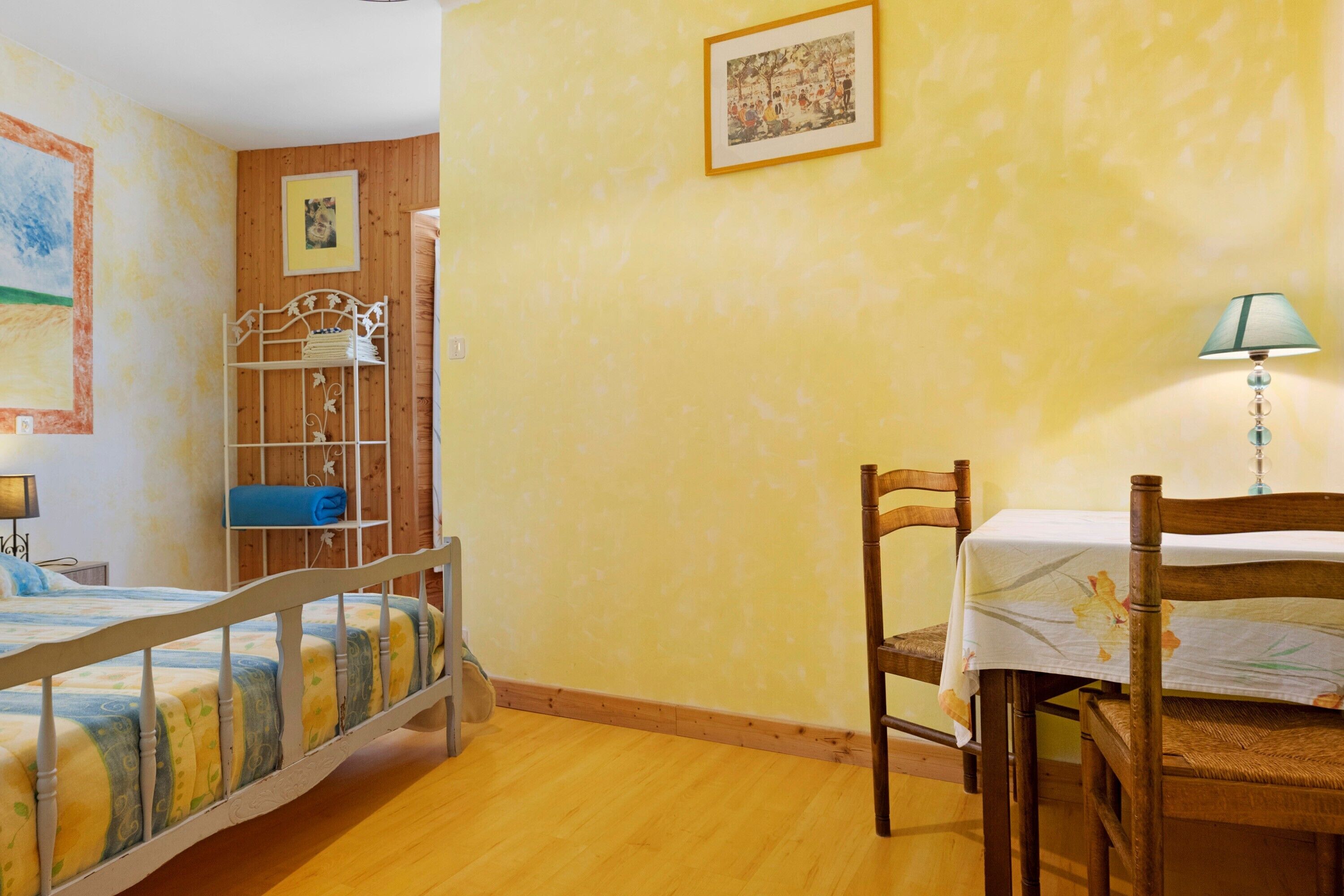 A room with a wooden floor, a bed with a white headboard, a wooden chair, a table with a tablecloth, a lamp, and a shelf with folded towels.