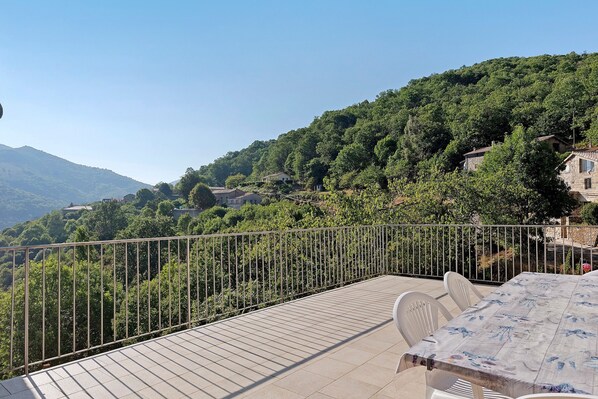 A terrace with a view of a mountainous landscape, a metal railing, and a table with chairs.