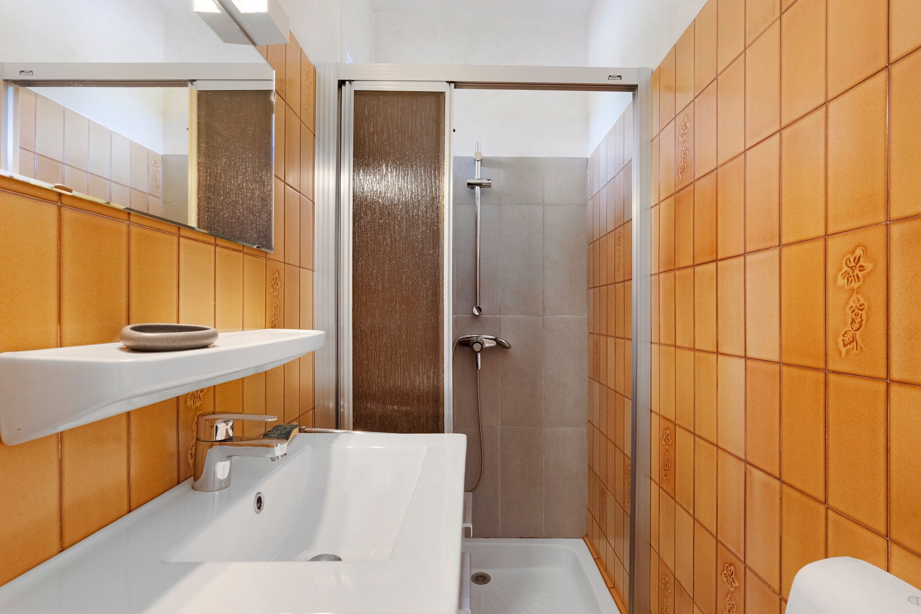 Une salle de bain avec un lavabo blanc, un miroir, une douche et des murs recouverts de carreaux orange.