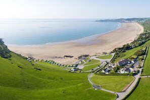 Vue aérienne d’une zone côtière avec une plage, des champs verdoyants et un ensemble de maisons et de caravanes.