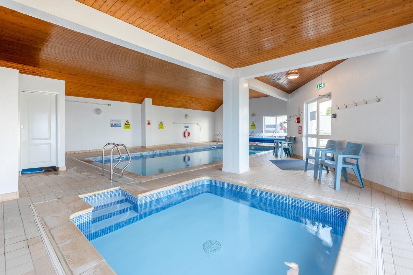 An indoor swimming pool with tiled flooring, wooden ceiling beams, and a seating area with chairs.