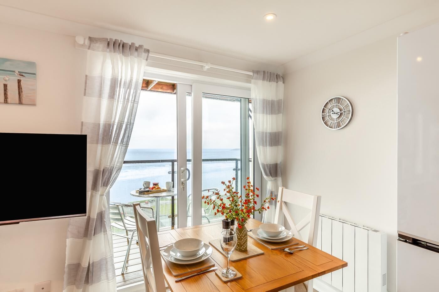 En matplats med ett bord dukat för två personer, utsikt över havet genom en glasdörr och en väggklocka.