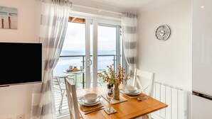 Et spiseområde med et bord dækket til to personer, udsigt til havet gennem en glasdør, og et vægur.