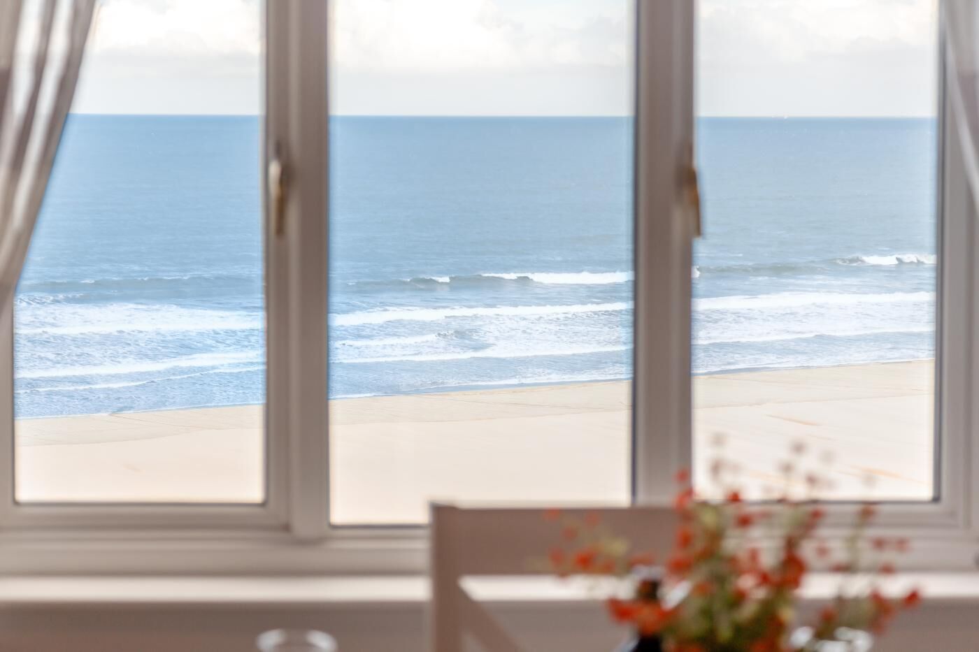 A view of the ocean through a window with a chair and a vase of flowers in the foreground.