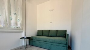 Et værelse med en grøn sofa, et lille sofabord, et vindue med gardin og en lyskontakt på væggen.