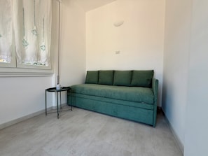 Ein Zimmer mit einem grünen Sofa, einem kleinen Beistelltisch, einem Fenster mit Vorhang und einem Lichtschalter an der Wand.