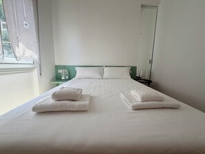 Ein ordentlich bezogenes Bett mit weißen Leinen und Handtüchern, ein grüner Kopfteil, ein Nachttisch mit einem Glas und ein Fenster mit einem weißen Vorhang.