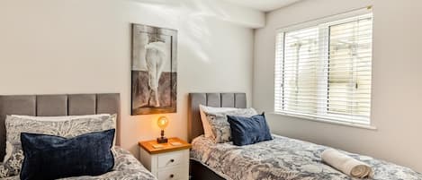 Un dormitorio con dos camas, una mesita de noche con lámpara, una ventana con persianas y un cuadro en la pared.