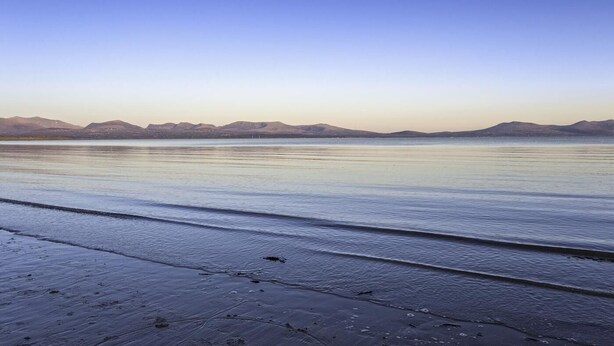 Una platja tranquil·la amb ones suaus, cel serè i muntanyes llunyanes.