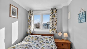 Ett sovrum med en säng, ett sängbord med en lampa, ett fönster med gardiner, en inramad tavla på väggen och en skylt med text.