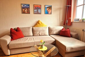Ein beiges Sofa mit roten und gelben Kissen, ein Glastisch mit einer Blume sowie drei gerahmte Bilder an der Wand.