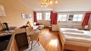 Een compacte hotelkamer met een kitchenette, een slaapbank, een bed en grote ramen voorzien van gordijnen.