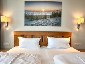 A hotel room with a wooden headboard, two white pillows, a beach scene painting, and wall-mounted lamps. - Moderne Ferienwohnung mit Balkon in Sellin (Sellin)