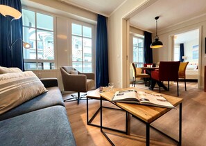 A modern hotel room with a sofa, a wooden coffee table with magazines, a dining area with chairs, and large windows with curtains. - Moderne Ferienwohnung mit Balkon in Sellin (Sellin)