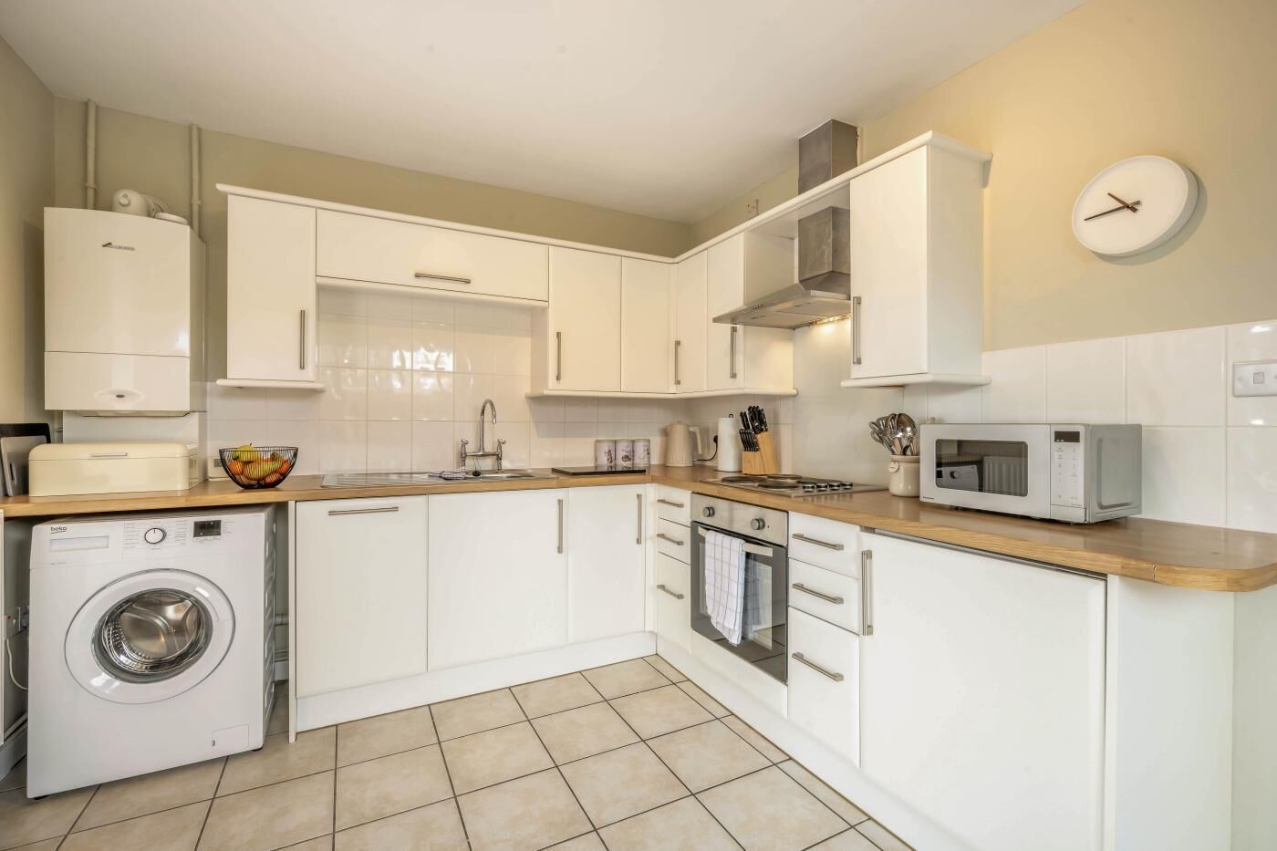 Una cocina compacta con muebles blancos, lavadora, microondas y un reloj en la pared.