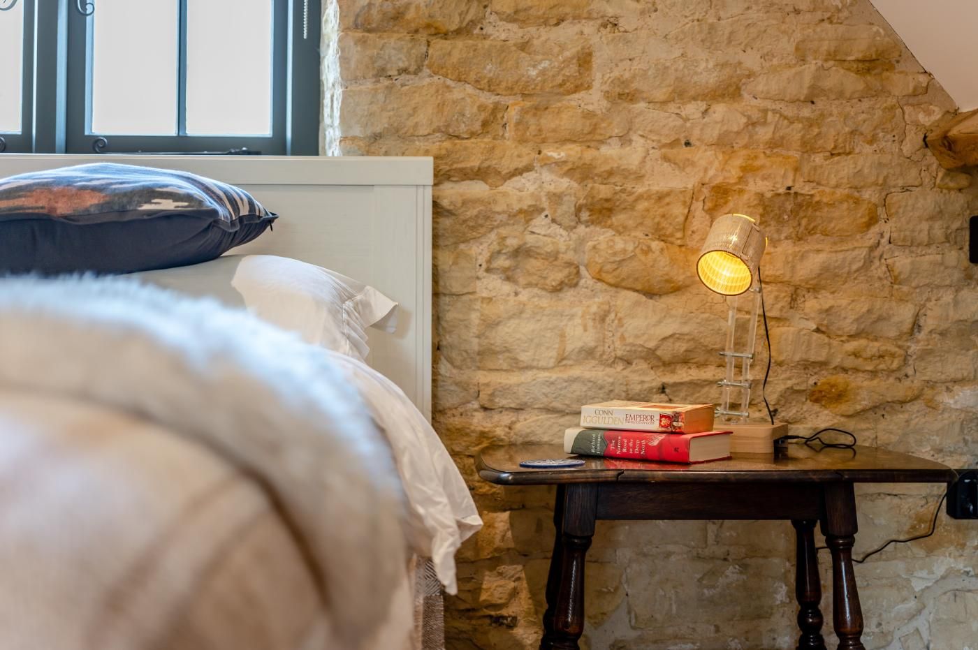 A bedroom with a stone wall, a bedside table with books, and a lamp.