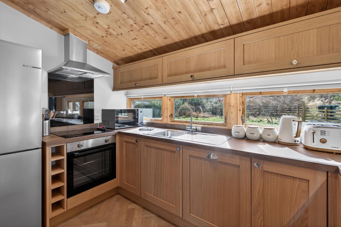 Eine moderne Küche mit Holzschränken, einem Edelstahl-Kühlschrank, einer Mikrowelle, einem Backofen und einem Fenster mit Blick ins Freie.